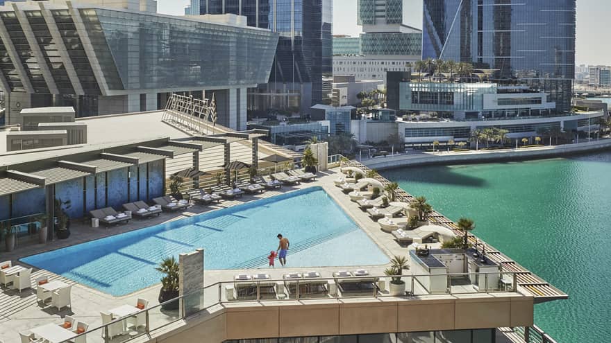 A wide shot of a man and child walking around a triangular pool overlooking the water and Abu Dhabi skyscrapers in the background. 