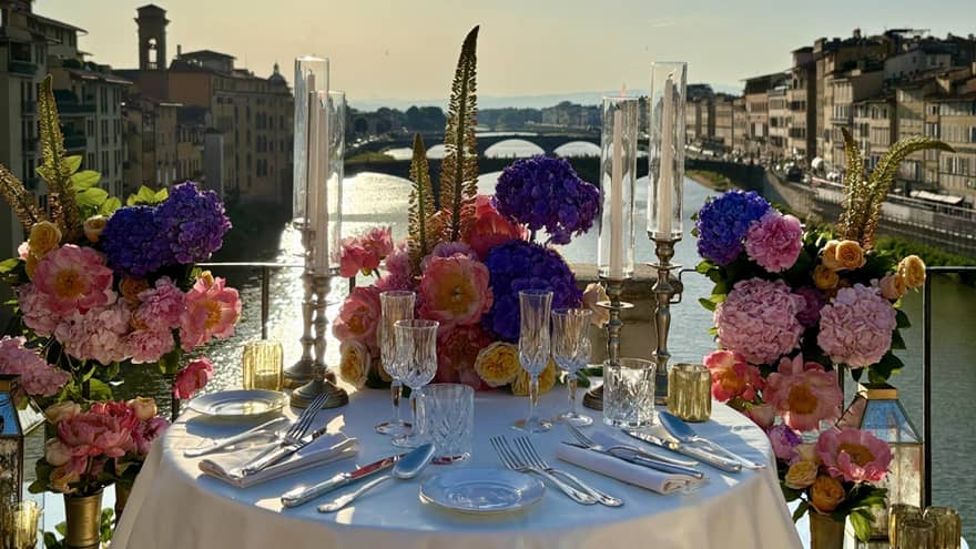 An elegantly set outdoor dining table overlooking a a river, with a sunny sky in the background.