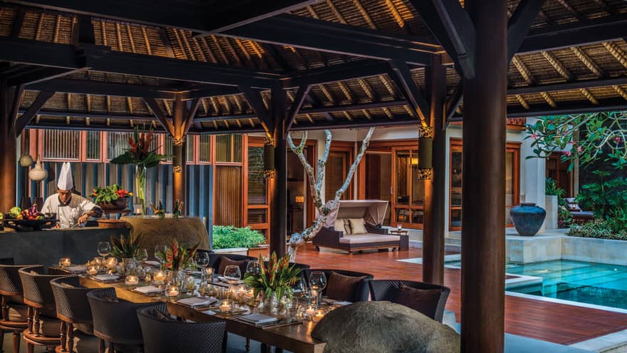 Chef prepares meal in front of elegant 10-person private dining table under thatched-roof gazebo, beside pool