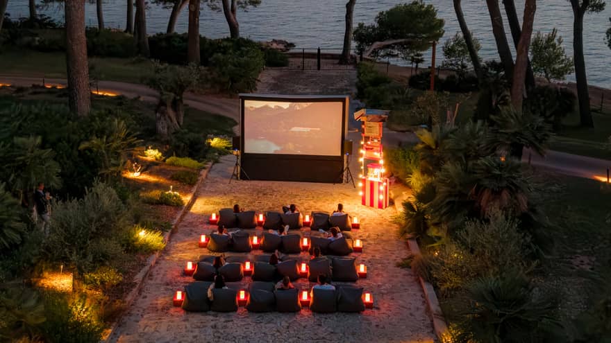 A bird’s-eye view of people seated in rows of chairs in front of a large movie screen in an outdoor setting in the evening, surrounded by trees, with the ocean in the background