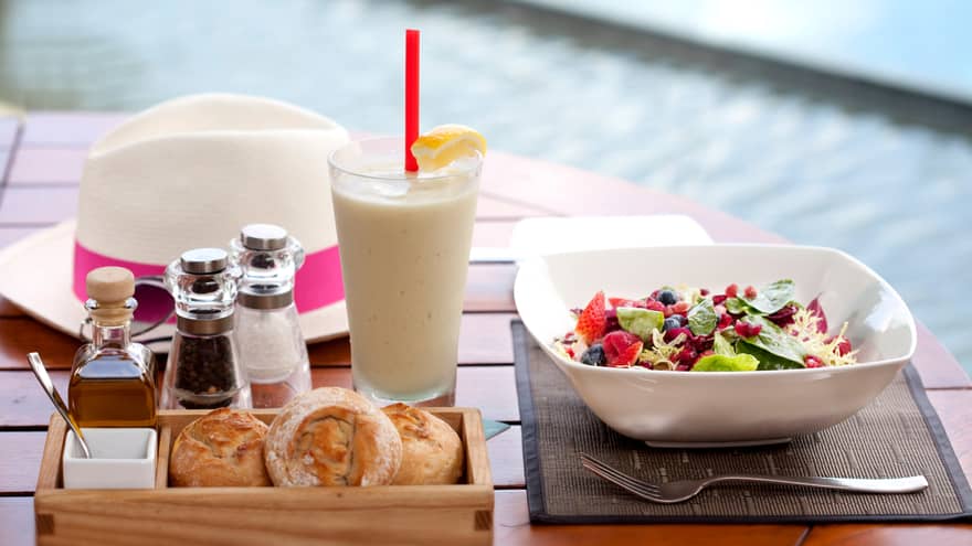 A table by the pool terrace with a meal of mixed salad, bread, condiments, and a cold beverage with lemon. A white hat with a pink band rests in the background.