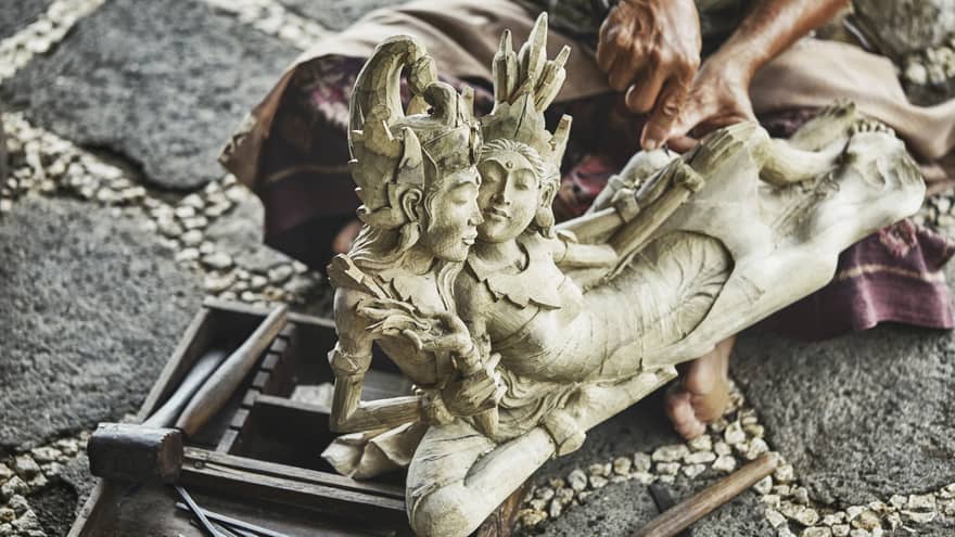 Close-up of intricate carved wood statue, man's hands with carving knife visible behind