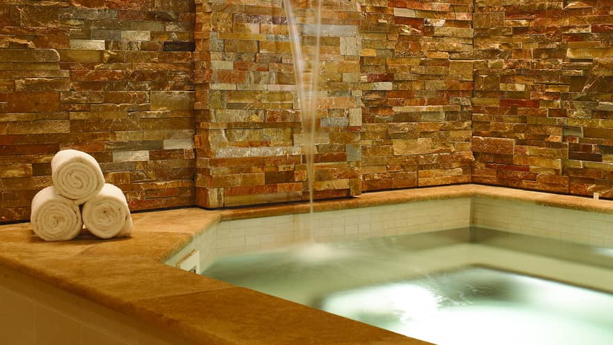 Glowing spa tub surrounded by stone wall and three rolled white towels on ledge