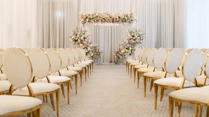 A stunningly decorated wedding aisle flanked by beautifully arranged cream and gold chairs, leading up to flower arch at altar, ready to welcome guests
