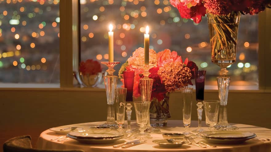 Honeymoon suite table setting with two long candles, pink flowers, city lights at night seen out window