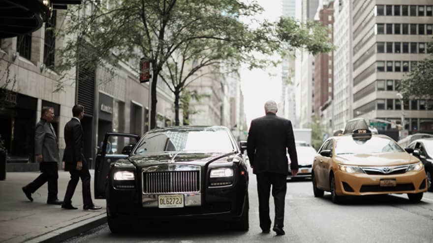 Back of man in suit, two men on sidewalk approaching black luxury car parked by curb