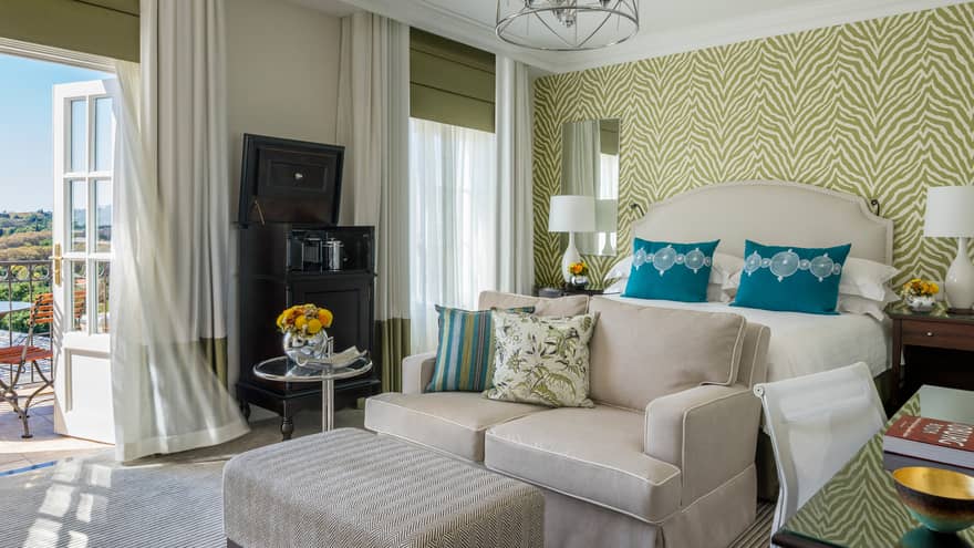 Panoramic View Deluxe Room white loveseat and ottoman against patterned wallpaper, open door to sunny patio