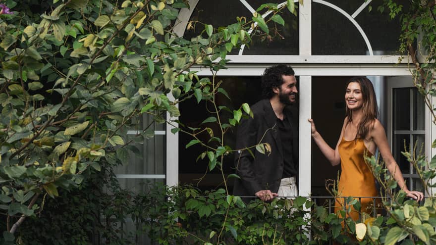 Two people stand on a vine-covered balcony surrounded by foliage.