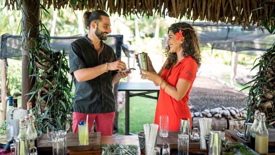 Two people hold a shaker and fresh herbs in a straw-roofed hut over a bar strewn with glasses, liquor bottles and garnishes. 