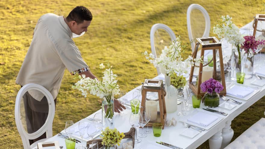 nHotel staff sets long outdoor wedding dining table with white flowers, lanterns