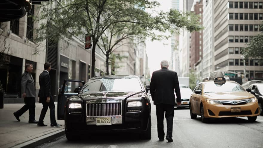 Back of man in suit, two men on sidewalk approaching black luxury car parked by curb