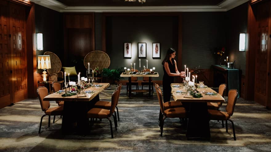 Intimate wedding dinner setup in a luxurious dining room at Four Seasons Whistler. The room features dark wood paneling and soft lighting, creating a warm and inviting atmosphere. Two wooden tables are elegantly set with white candles, floral arrangements, and fine dinnerware. A woman in a dark dress adjusts the table settings, preparing for the event. The sophisticated decor and thoughtful arrangement reflect a serene and exclusive wedding venue.,Intimate wedding dinner setup in a luxurious dining room at Four Seasons Whistler. The room features dark wood paneling and soft lighting, creating a warm and inviting atmosphere. Two wooden tables are elegantly set with white candles, floral arrangements, and fine dinnerware. A woman in a dark dress adjusts the table settings, preparing for the event. The sophisticated decor and thoughtful arrangement reflect a serene and exclusive wedding venue.