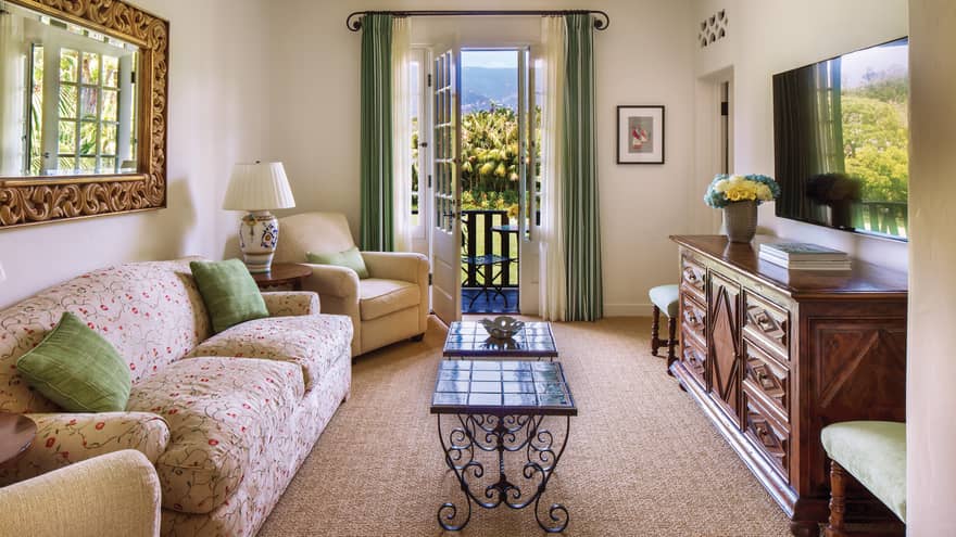 Floral loveseat with green accent pillows, across from wood dresser, open balcony door