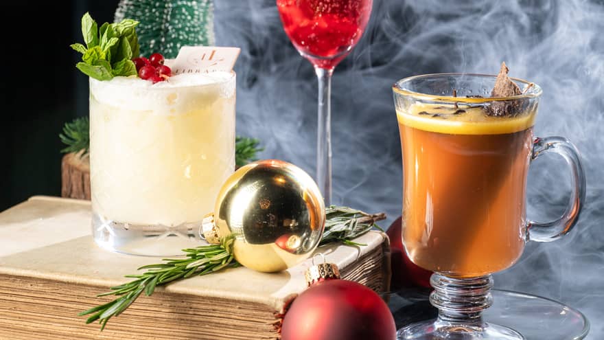 Three festive cocktails arranged on a table with gold and red ornaments, small trees, an old book and a puff of smoke in the background