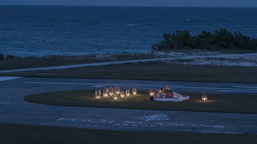 Two people stargaze by ocean at night on lounge chairs surrounded by glowing lanterns