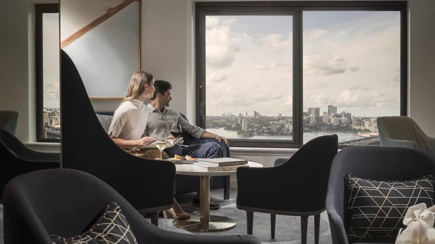 Couple enjoy wine and cheese in hotel room seating room by window overlooking Sydney