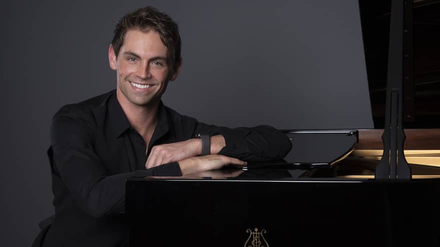 Person wearing black button-down collared shirt and black pants sits at a Steinway 
