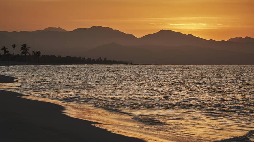 Beach and ocean in Los Cabos, Mexico, with sun setting behind distant mountains.