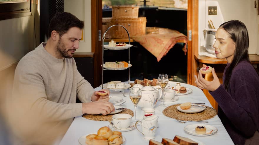 Two people sit at a dining table in a cozy indoor dining room. The table is set with plates of food, and a cottage-like interior with windows showing greenery outside is in the background.