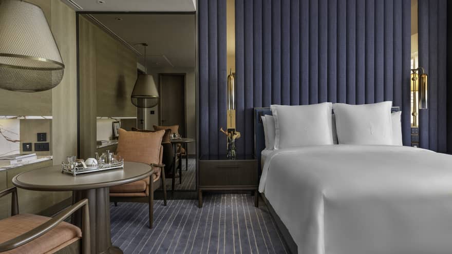Renovated Superior Room with queen bed and white linens, blue striped carpet and accent wall, floor-to-ceiling mirror, table for two with salmon armchairs