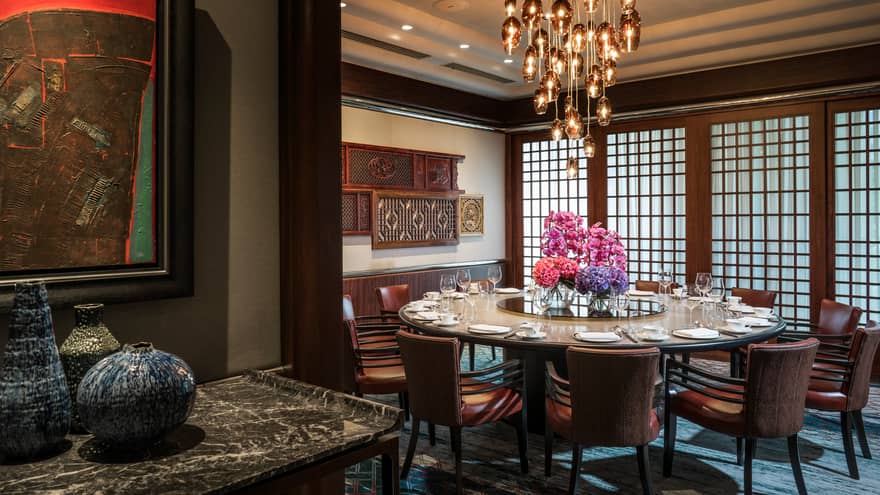 Round private dining table with pink flowers under modern hanging lights, wood carvings on wall