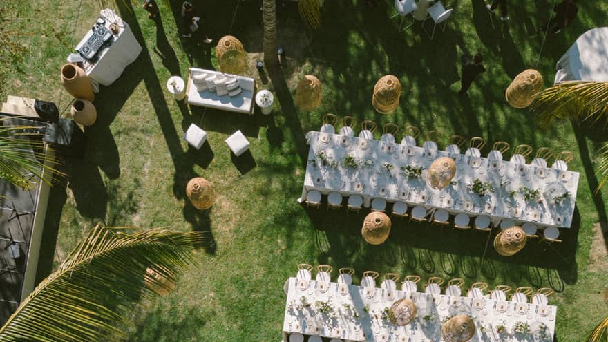 An aerial view of an outdoor event setup on a green lawn surrounded by palm trees. Setup includes long dining tables set for an event, chairs, lighting and other decor.