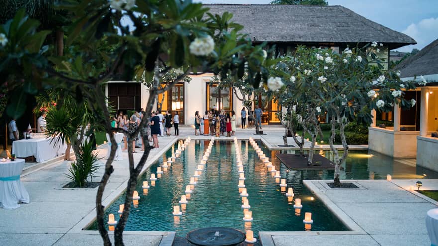 View past tropical tree to guests at patio reception around pool with floating candles