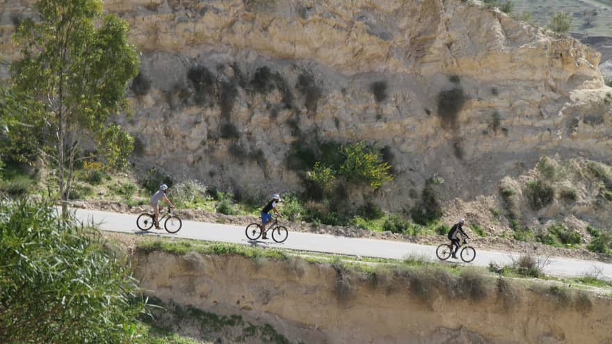 Side view of cyclists riding their bicycles down a paved road surrounded by trees and rocky cliffs on a sunny day.