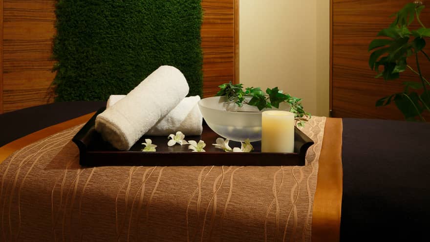 Wood tray with rolled white towels, flowers, candle, bowl with vine on spa bed