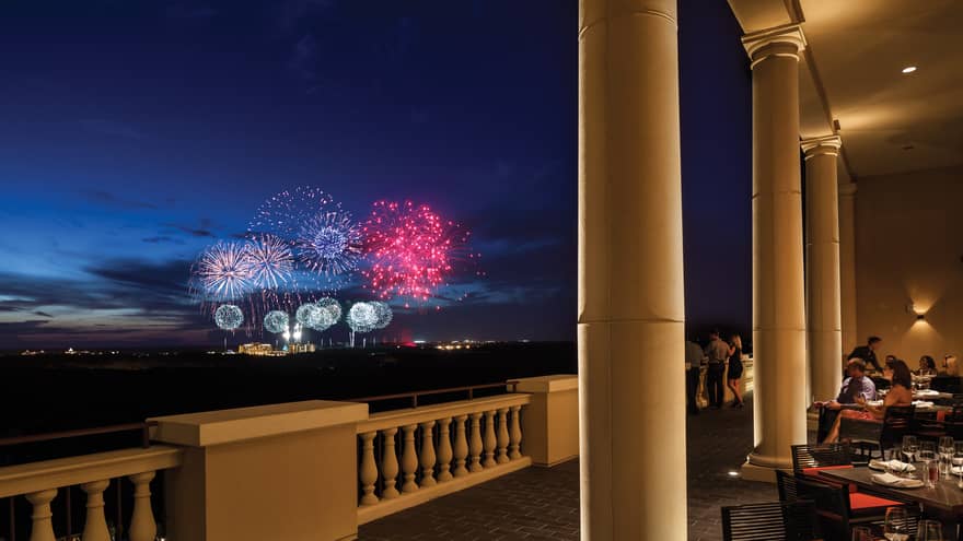 The terrace of a restaurant with tables and people watching fireworks in the distance.