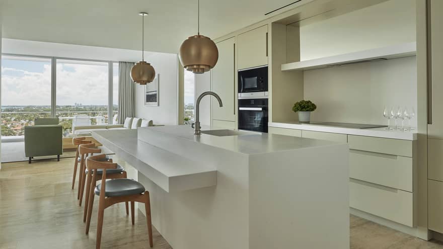 Retro-style brass lamps above white kitchen island with sink and stools. This kitchen is part of the private residences at Four Seasons Hotel at the Surf Club, Surfside, where luxury vacation rentals in Miami are luxuriously situated.