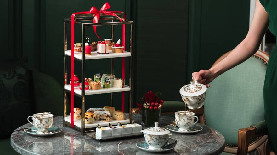 A round grey marble table is set with three tea cups and a three-tiered serving platter filled with pastries and wrapped in a red bow