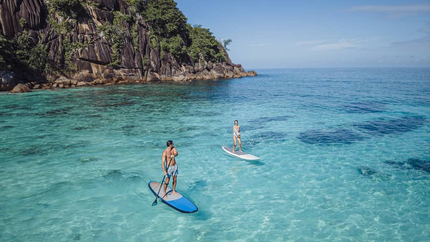 Two people stand-up paddleboarding on clear blue water with rocky cliff in back drop