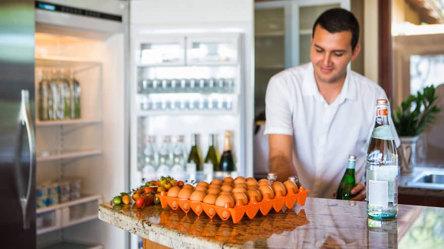 Smiling man holds green bottle in front of open fridge, fresh eggs on counter