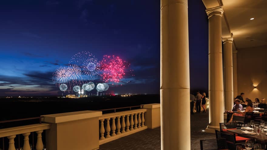 The terrace of a restaurant with tables and people watching fireworks in the distance.