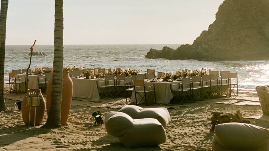 Dining tables for an event set near palm trees on the beach on a sun-drenched day, with a dramatic cliff in the background