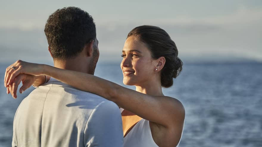 Couple stands oceanside at sunset, her arms around his neck
