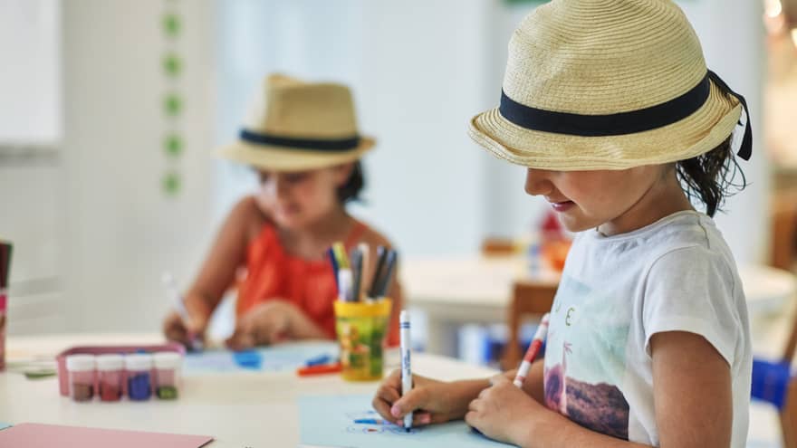 Two smiling children wearing straw fedora hats colour pictures with markers at craft table