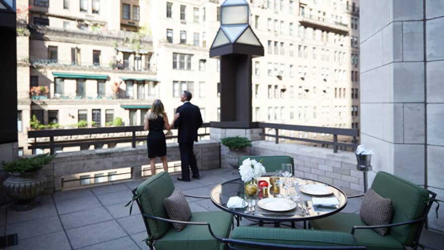 Man and woman in business suits look out from rooftop patio with table, plush chairs