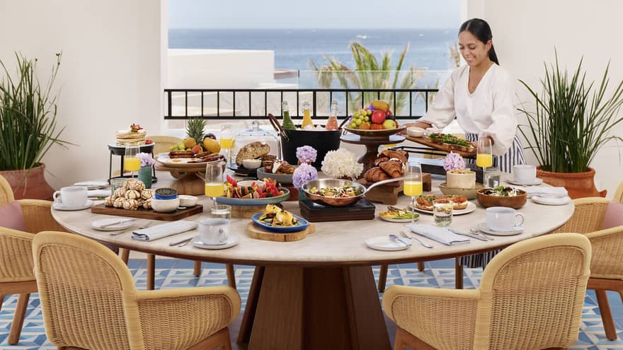 Woman sets a large round table with ocean view, with a cluster of rattan lights hanging above and blue tiled floor
