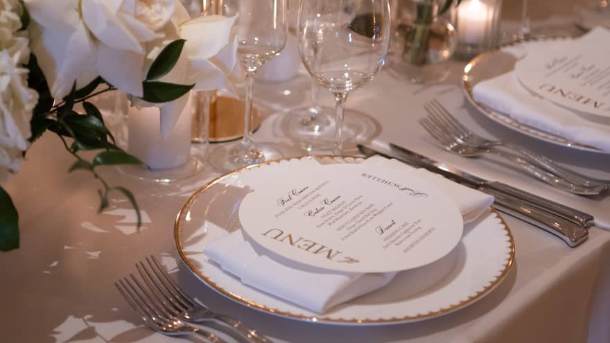 A dining table set with wine glasses, candles, a menu and bouquets of flowers