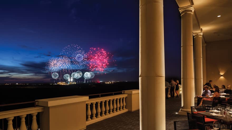 The terrace of a restaurant with tables and people watching fireworks in the distance.