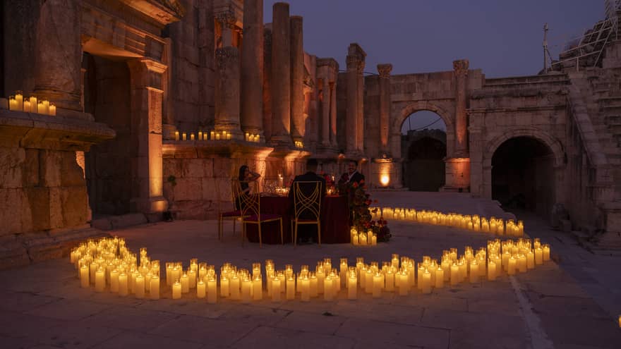 A circle of lit candles surrounds a small dining table set in the middle of an ancient site featuring stone columns and archways with the night sky as a backdrop