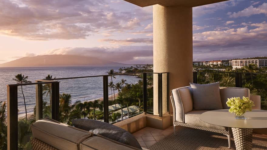 Suite balcony with cushioned sofas and a table overlooking the ocean and palm trees at sunset