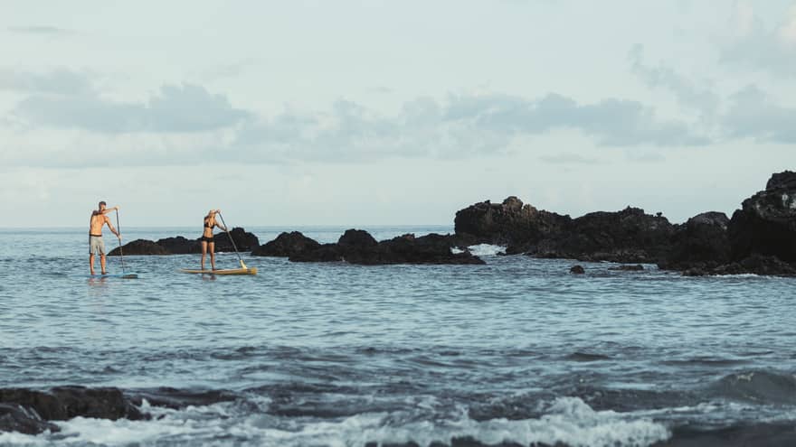 Two people balance on stand-up paddleboards in lagoon by black rocks