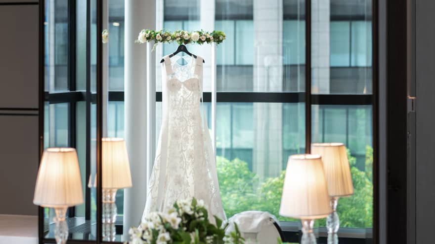 Reflection in a tall mirror of a wedding gown hanging across a window in a room with chandelier and white floral décor.