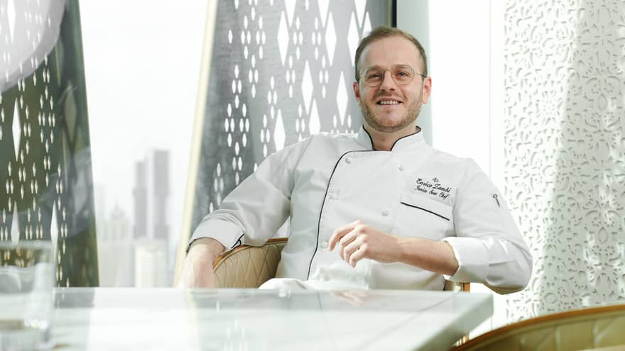 Chef with short light-brown hair, slight beard and wearing white chef's jacket sits back in a cream-coloured club chair at a restaurant table with a city skyline in the background