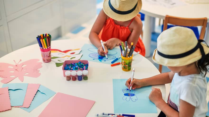 Two young girls wearing straw sun hats colour pictures at craft tables