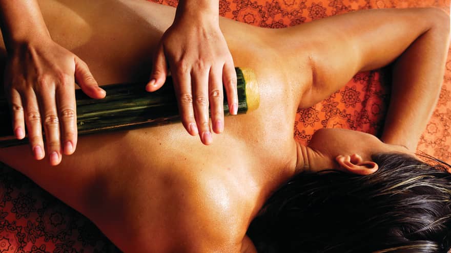 Spa staff rolls thick bamboo stick over woman's bare back, shoulders 