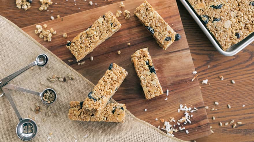 Five granola bars are scattered on a wooden table with a tray of baked granola, a burlap table tunner and stainless steel measuring spoons laying nearby